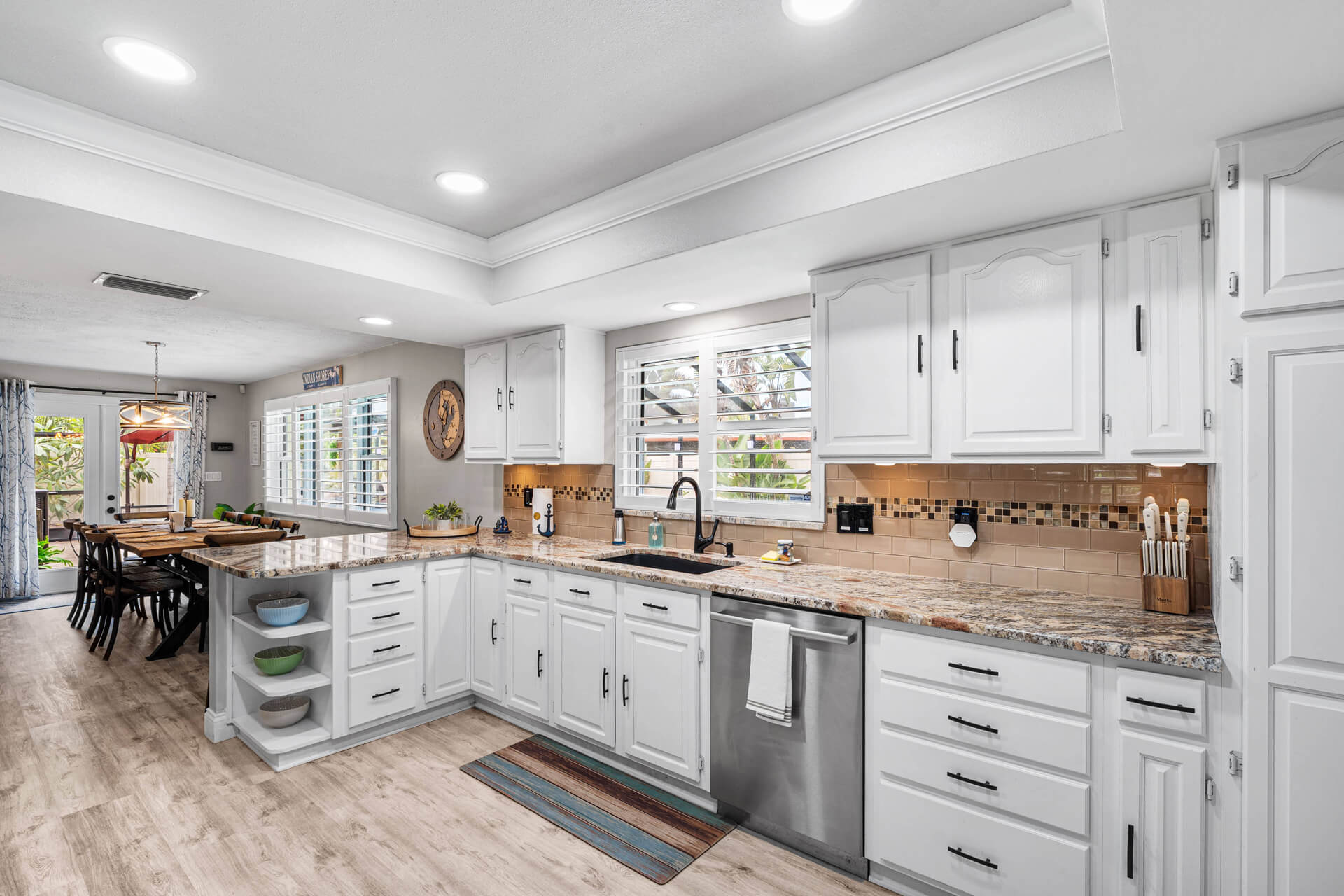 A fully equipped kitchen with granite countertops, white shaker cabinets, stainless steel dishwasher, and a view into the sunlit dining room — everything a large group needs for gourmet meals at Twin Oaks.