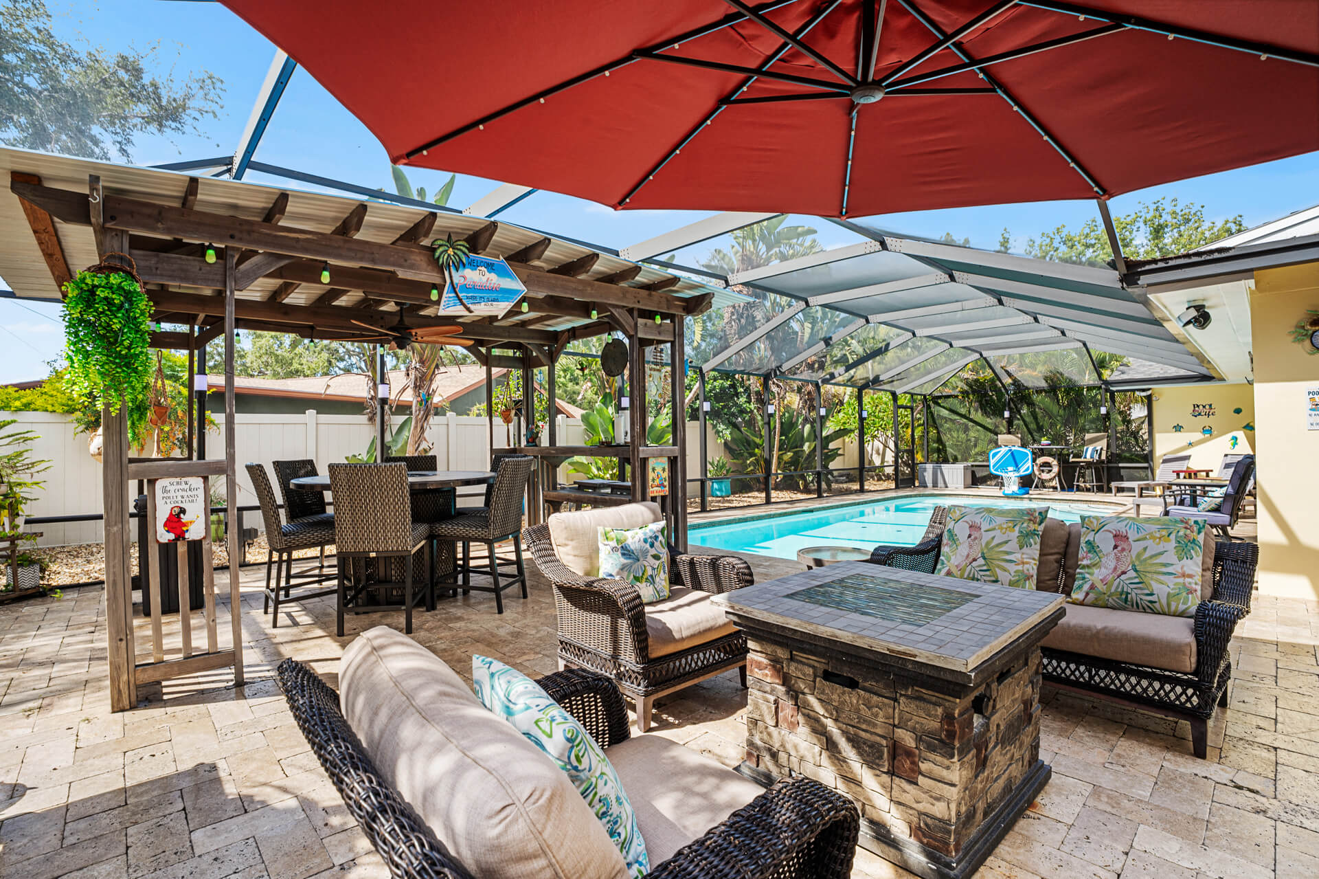 The heart of the backyard: a covered Tiki Bar with high-top seating, wicker lounge furniture, and a stone fire pit table — all steps from the pool at Twin Oaks Coastal Luxury.