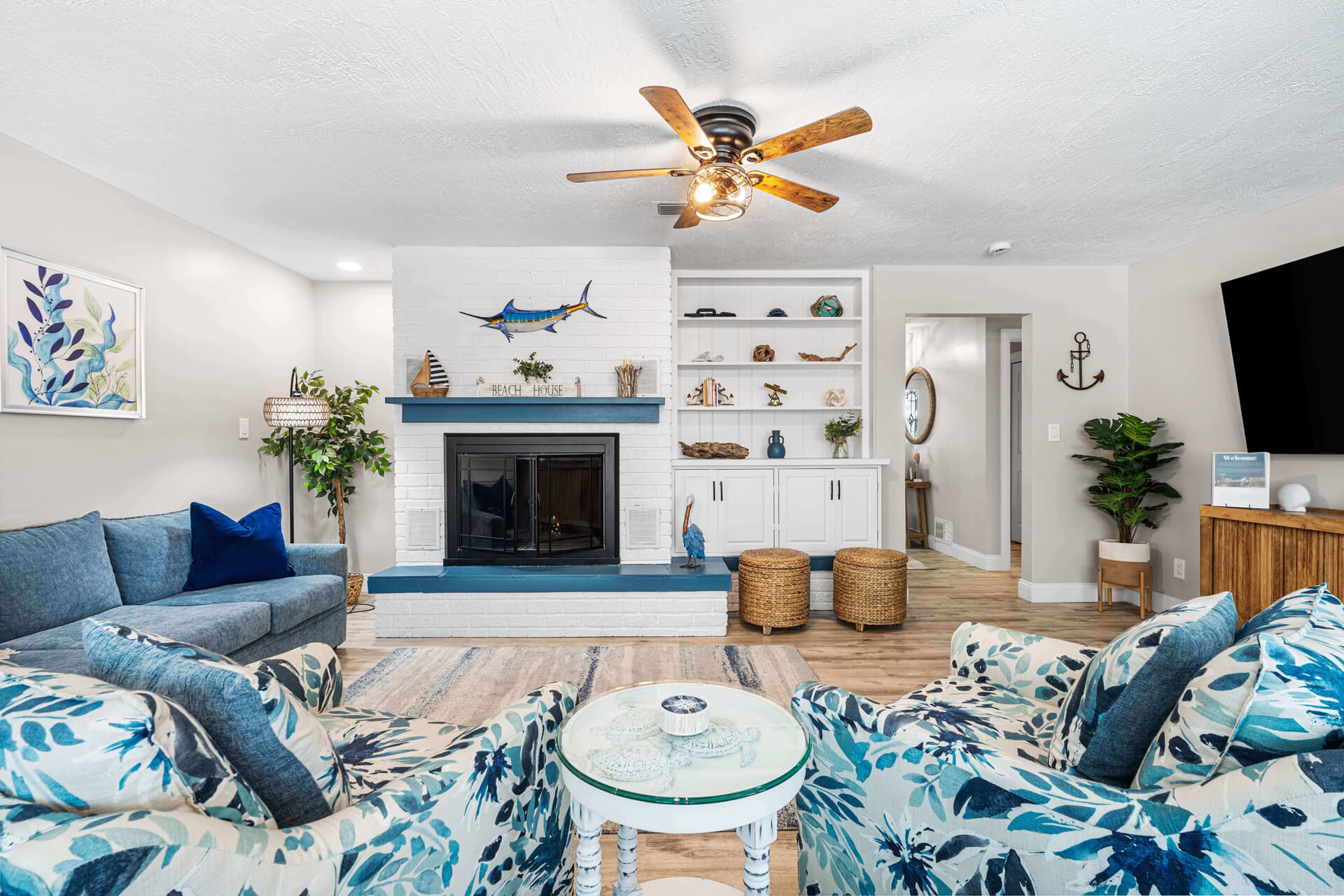 A beautifully appointed coastal living room featuring a white brick fireplace with blue accents, a marlin wall sculpture, built-in shelving, and plush nautical-themed seating at Twin Oaks Coastal Luxury.