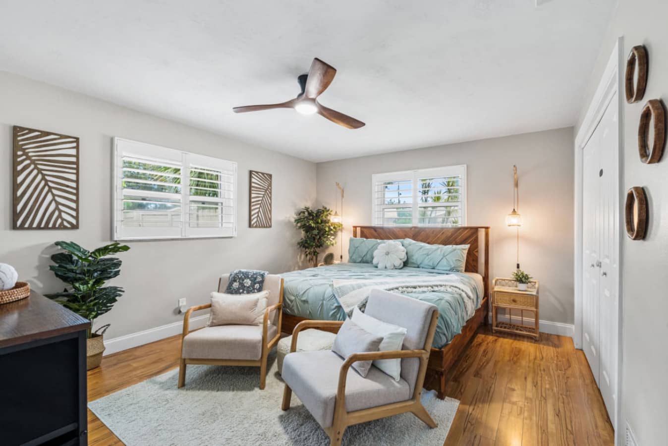 Palm Paradise, the primary suite at Twin Oaks Coastal Luxury, features a king bed with soft seafoam linens, warm pendant lighting, hardwood floors, and a private ensuite with rain shower.