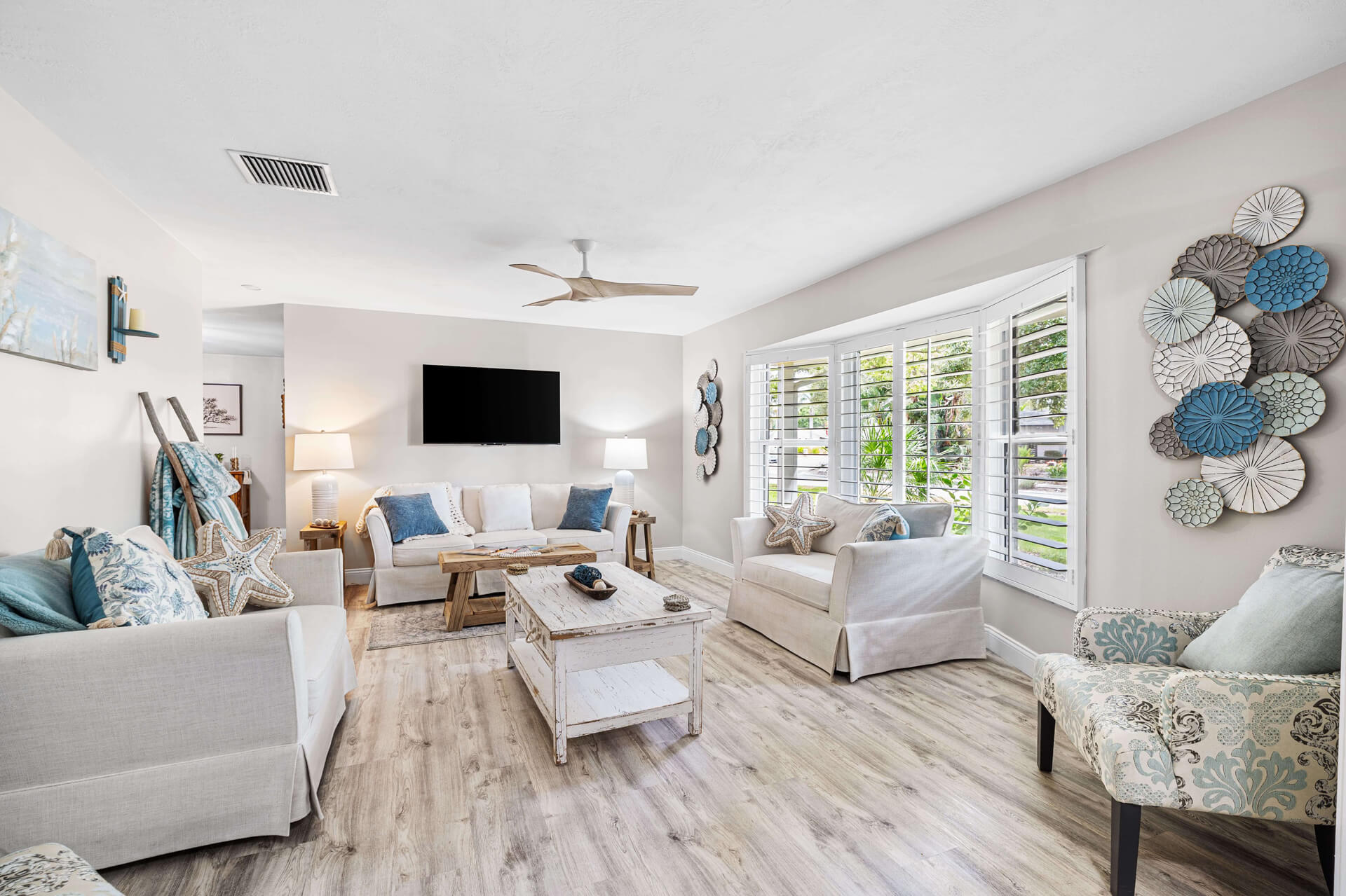 A bright, airy second living space with white slipcovered sofas, a distressed wood coffee table, starfish accents, and large plantation shuttered windows overlooking lush Florida greenery.