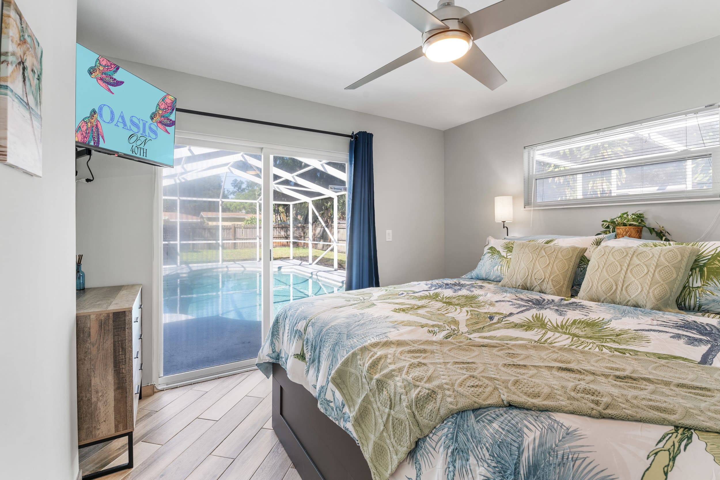 Primary bedroom at Sun Oasis — king bed with tropical palm-print linens, wall-mounted smart TV, and direct sliding glass door access to the private screened pool.